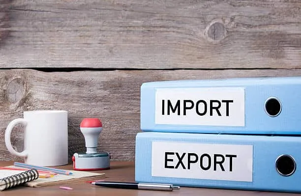 Folders labeled 'IMPORT' and 'EXPORT' on a desk with a stamp and paperwork, representing the organization of documents required for international trade.