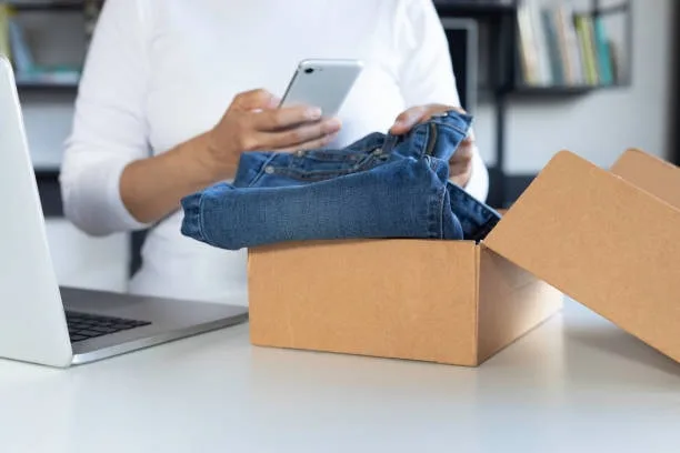 A person packing jeans into a shipping box from a home office, using a smartphone to coordinate the sale, demonstrating the online retail process for clothing.