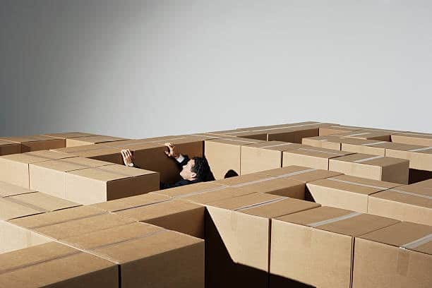 Man lost in a maze of cardboard boxes, symbolizing confusion and delays caused by packages stuck in customs.