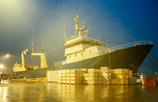 Cargo vessel docked at night with stacked heavy freight crates at port.