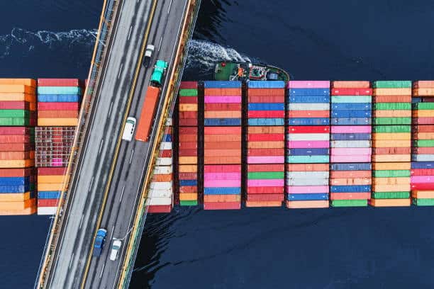 Aerial view of a container ship passing under a bridge with trucks, representing multimodal transport in outbound logistics operations.