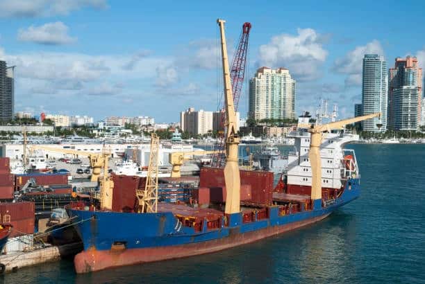 Heavy cargo container ship docked near urban port with cranes unloading freight.