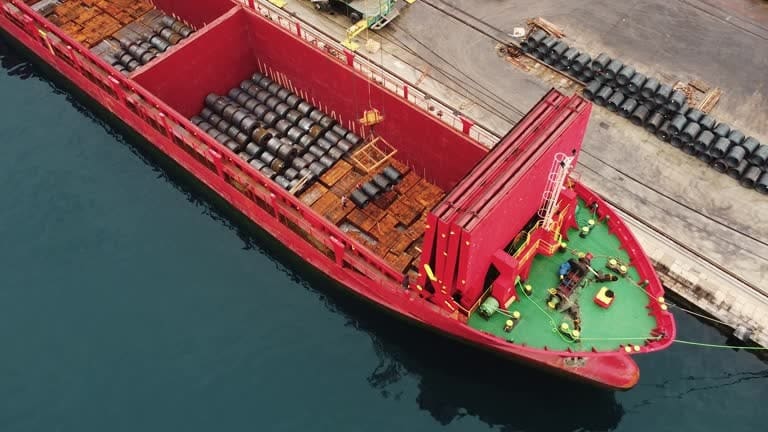 Top-down view of red cargo vessel transporting heavy industrial barrels by sea.