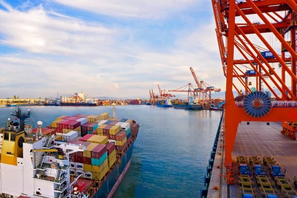 A cargo ship docked at a port with colorful shipping containers and red cranes in the background, under a partly cloudy sky.