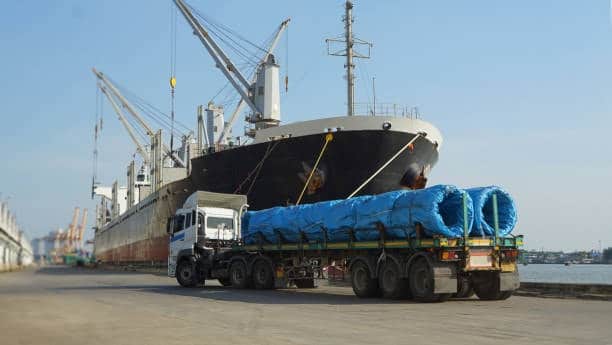 Truck carrying heavy coiled cargo to be loaded onto a docked ship.