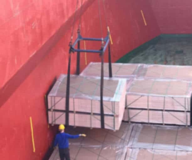Dock worker guiding large palletized cargo into ship hold using crane straps.