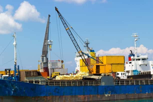 Crane loading heavy cargo and containers onto a shipping vessel at port.