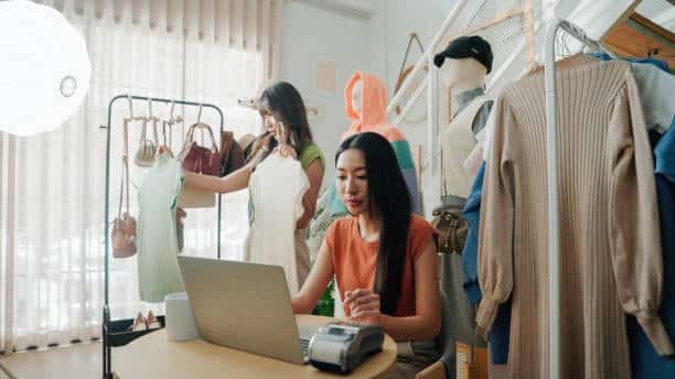 A boutique staff member processes a transaction on her laptop while customers browse clothing racks in the background, illustrating the daily operations of a small fashion business.