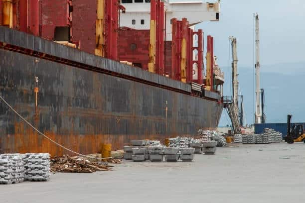 Heavy metal ingots stacked near a rust-colored cargo ship at export dock