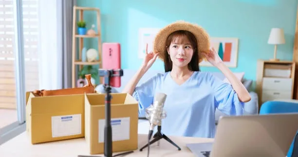 A cheerful fashion vlogger adjusting her straw hat while sitting at a desk with a microphone and shipping boxes, ready to start a live session promoting products.