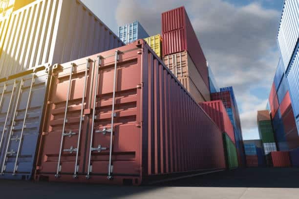 Multiple shipping containers stacked at a freight terminal, representing storage and staging for door delivery logistics.