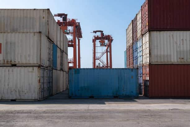 Shipping containers stacked at a port terminal with cranes in the background, showing the starting point of port-to-door logistics.
